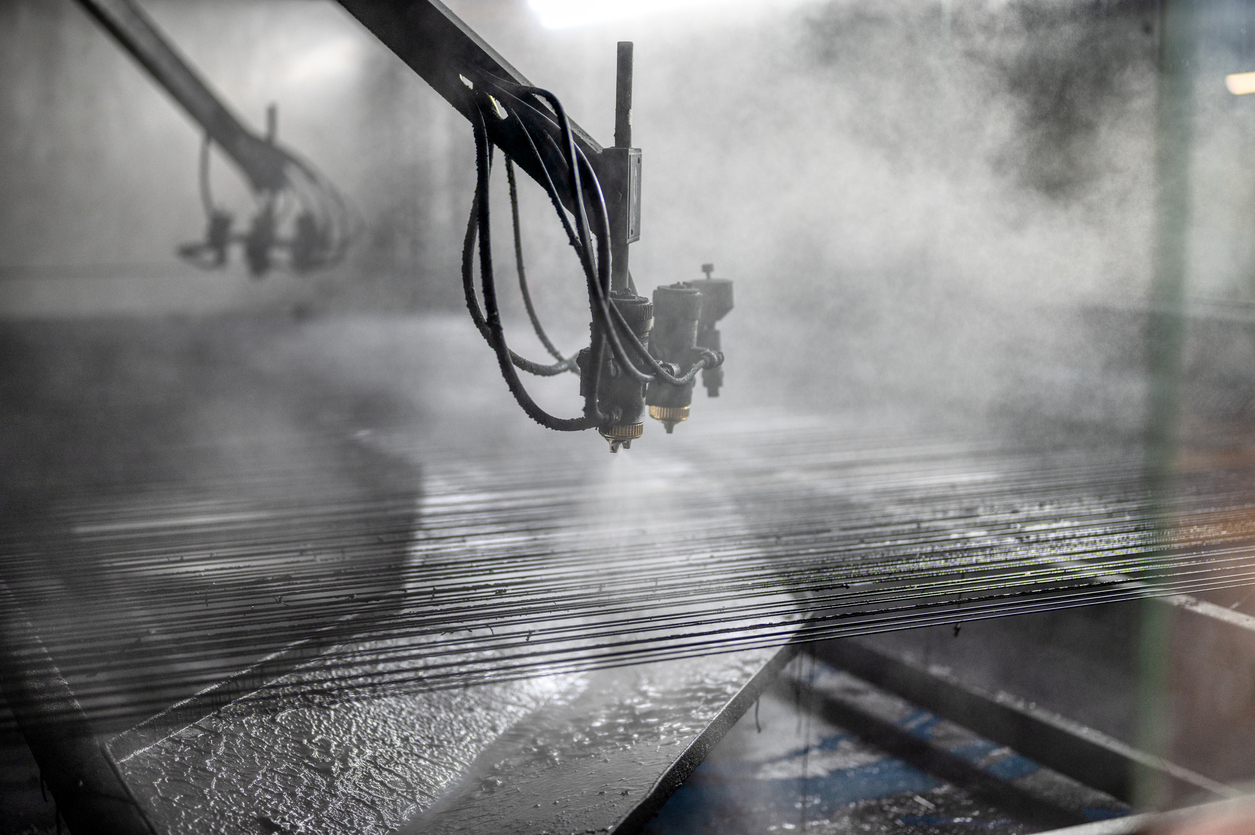 Industrieroboterarme mit Düsen oder Werkzeugen bewegen sich über parallel angeordnete Metallstäbe, aus denen Dampf oder Nebel aufsteigt. Die Szene wirkt technisch und ist von feuchter, dampfgesättigter Luft geprägt.