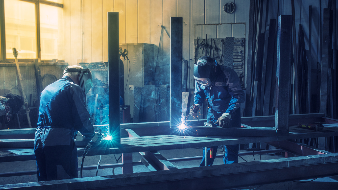 Zwei Personen in Schutzkleidung schweißen Metallrahmen in einer Werkstatt mit großen Fenstern