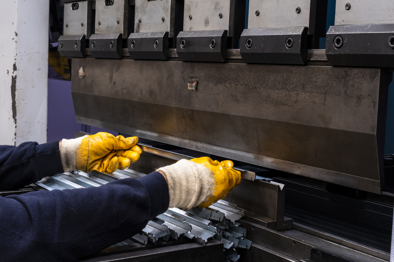 Eine Person mit gelben Schutzhandschuhen hält ein längliches Metallstück an eine große Industriemaschine. Die Maschine hat mehrere gleichmäßig angeordnete, senkrechte Metallteile an der Oberseite. Im Vordergrund liegen weitere Metallprofile, die parallel zueinander ausgerichtet sind. Die Szene zeigt einen Arbeitsvorgang in einer Werkhalle.
