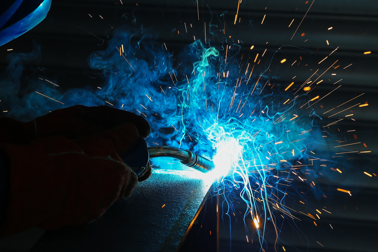 Ein Schweißvorgang mit intensivem blauen Licht und Funkenflug steht im Mittelpunkt. Eine Hand mit Schutzhandschuh hält das Schweißgerät an ein Metallstück. Dichte Rauchschwaden und leuchtende Funken breiten sich nach rechts aus. Im Hintergrund sind horizontale, unscharfe Strukturen zu erkennen.